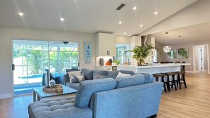 A spacious living room with a large sectional sofa, a dining area with wooden tables, and a kitchen with white cabinets. - SunCoast Luxury Estates-New, Spacious,  Large Heated Pool, 10 min to the Beach  (Venice)
