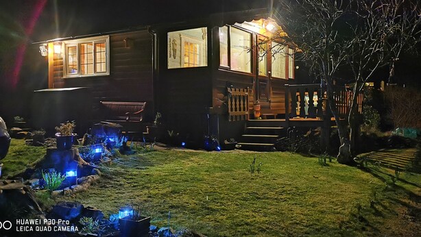 A wooden house with a porch, illuminated windows, and a small garden with blue lights. - Log Cabin - Free Parking, Garden & pet Friendly (Blaenau Ffestiniog)