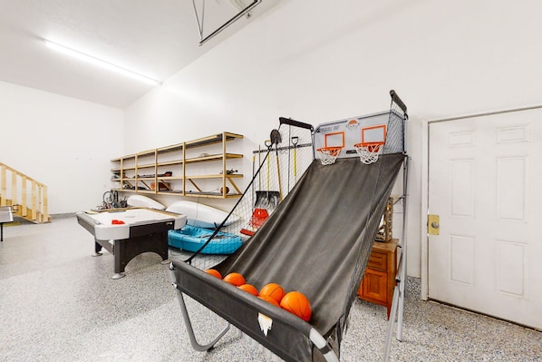 A room with a pool table, a basketball hoop, shelves with sports equipment, and a door.