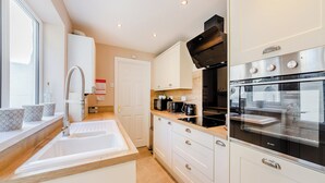 A modern kitchen with a sink, stove, and oven. There is a window with blinds, a door, and a countertop with a kettle and coffee maker.
