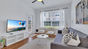 A modern living room with a flat-screen TV, a ceiling fan, wicker chairs, a gray sofa, and a large window with blinds. - Newly Renovated First Floor 2Bed & 2 Bath, Near Disney (Kissimmee)