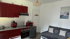 A compact kitchen with red cabinets, a microwave, a coffee maker, and a dishwasher. There is a gray sofa with decorative pillows and a painting of a landscape on the wall. - LES COMBES apartment-chalet sleeps 4 classified 3 * LES ROUSSES (Les Rousses)