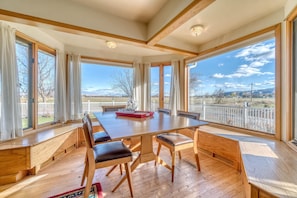A spacious dining area with a long wooden table, cushioned benches, and large windows offering a view of a fenced outdoor area. - Shady Acres: Two Living Spaces & Curated Library (Fruita)
