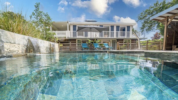 Ein klares Schwimmbecken mit einer Holzterrasse und Liegestühlen, einem Wasserfall aus Stein und einem Gebäude mit Balkon im Hintergrund.