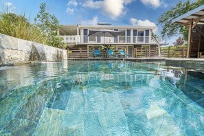 Una piscina cristalina con cubierta de madera y sillones de descanso, una cascada de piedra y un edificio con balcón al fondo.