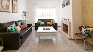 A living room with a brown leather sofa, a white coffee table, and a wicker chair. There is a fireplace with a brick surround and a white mantel. - Paradise Place - Pool Balcony (Nazaré)