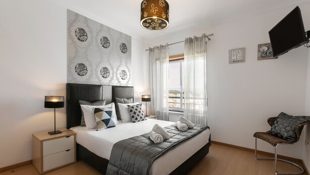 A modern bedroom with a large bed, bedside tables, a chair, and a TV mounted on the wall. - Paradise Place - Pool Balcony (Nazaré)