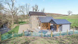 A stone house with a large porch and a small attached shed, surrounded by a fenced garden and a river.