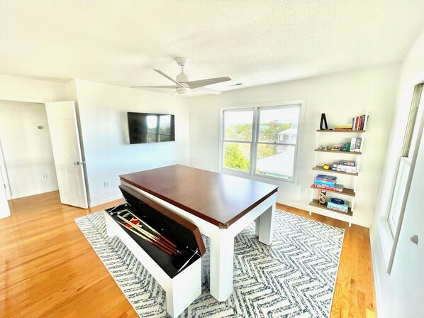 Una habitación amplia con una gran mesa de madera, un ventilador de techo, una estantería y una ventana con vistas al verdor.