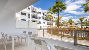 Een balkon met een witte tafel en stoelen, uitkijkend op een zwembad met palmbomen en een houten omheining.