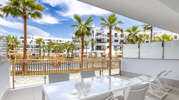 Een balkon met een glazen tafel en stoelen, met uitzicht op een zwembad en palmbomen, en een zicht op moderne gebouwen in de verte.