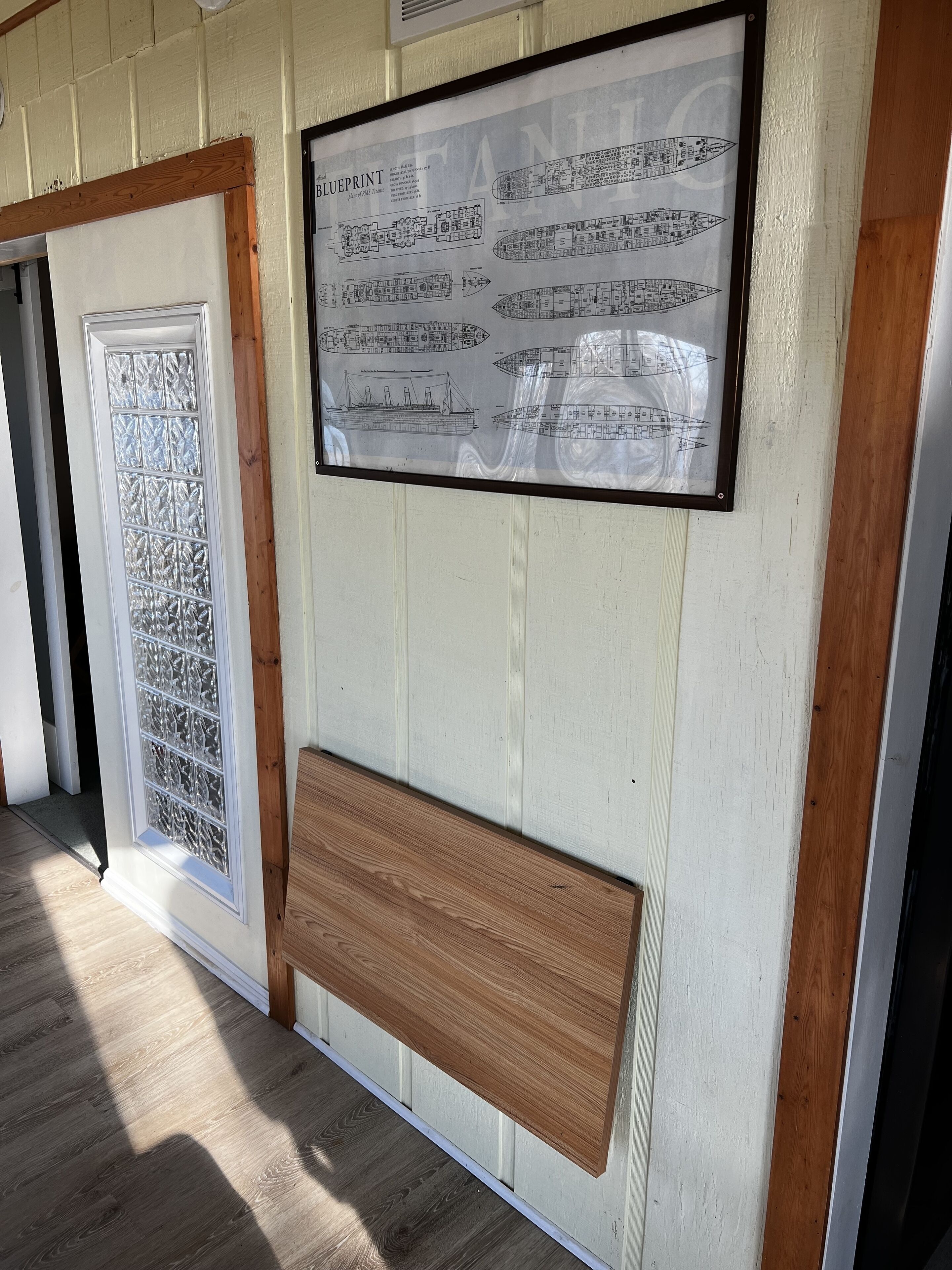 Een ingelijste blauwdruk van een scheepsinterieur, een decoratieve glazen deur en een houten bank.