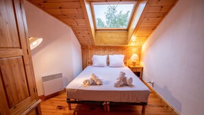 A bedroom with a sloped ceiling, wooden walls, a bed with white linens, two pillows, a bedside lamp, and a window with a view of trees.