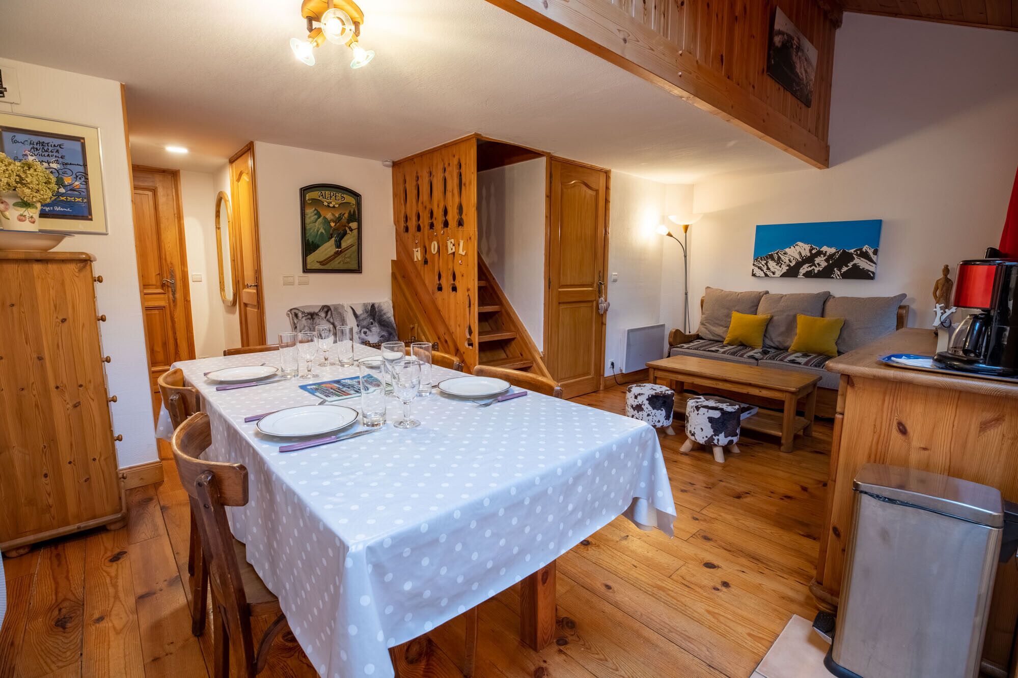 A dining area with a table set for four, wooden cabinets, a staircase, and a sofa in the background.