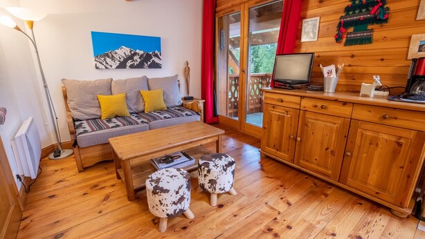 A cozy living room with a sofa, coffee table, and two ottomans. A television is on a wooden cabinet, and there's a painting of mountains on the wall.