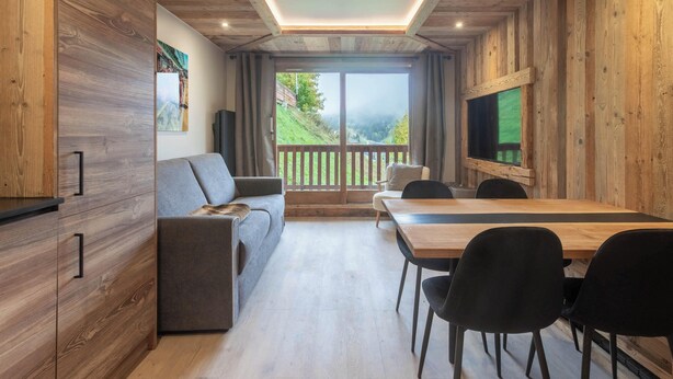 A modern interior with a wooden wall, a sofa, a dining table with chairs, and a large window with a view of a green landscape. - Appartement ski in and out in the center of Méribel (Les Allues)