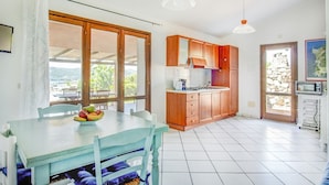 Une cuisine avec des meubles de rangement en bois, une table à manger sur laquelle se trouve un panier de fruits, et une vue à travers la porte vitrée.