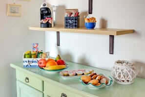 A kitchen counter with a coffee maker, a box of coffee pods, a bowl of bread, a fruit bowl, a plate of cookies, and a decorative lantern. - Palau Port-Maddalena - APT - Home and More (Palau)
