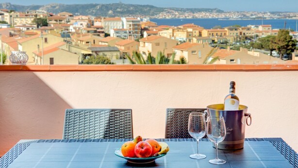 Een tafel met een fruitschaal, twee wijnglazen en een champagnefles op een balkon met uitzicht op een kustplaats.