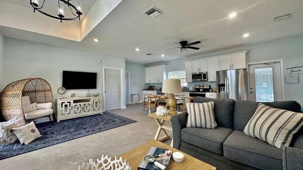 A spacious living room with a grey sofa, a television, and a decorative chair. A kitchen is visible in the background with white cabinets and a dining area. - Modern Beach Cottage - Just Steps to the Ocean and Down Town Cocoa Beach (Cocoa Beach)