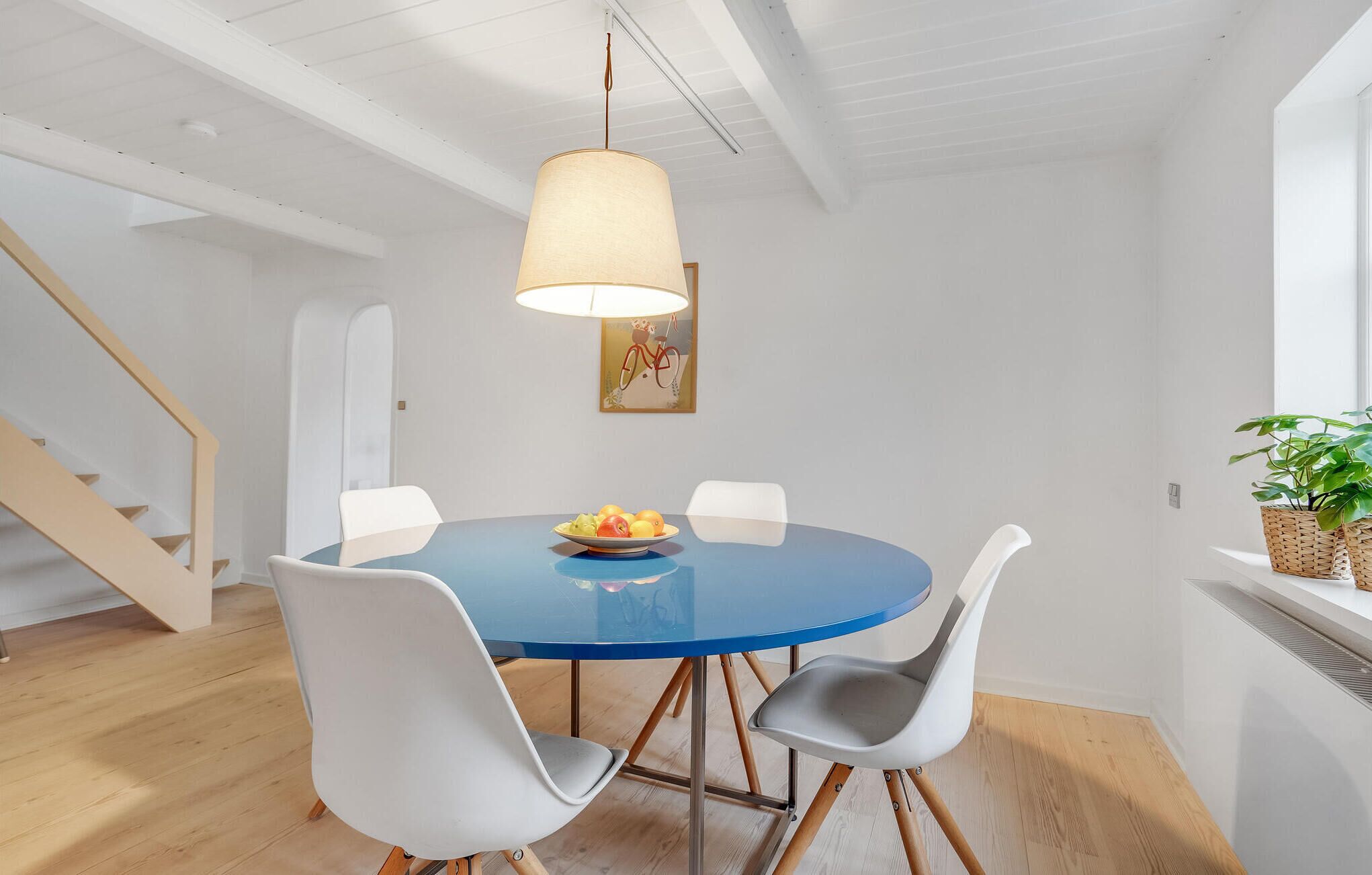 A dining area with a round table, four chairs, a hanging light fixture, and a staircase in the background.