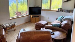 Een woonkamer met een bank, een stoel, een televisie en een raam met uitzicht op bomen.