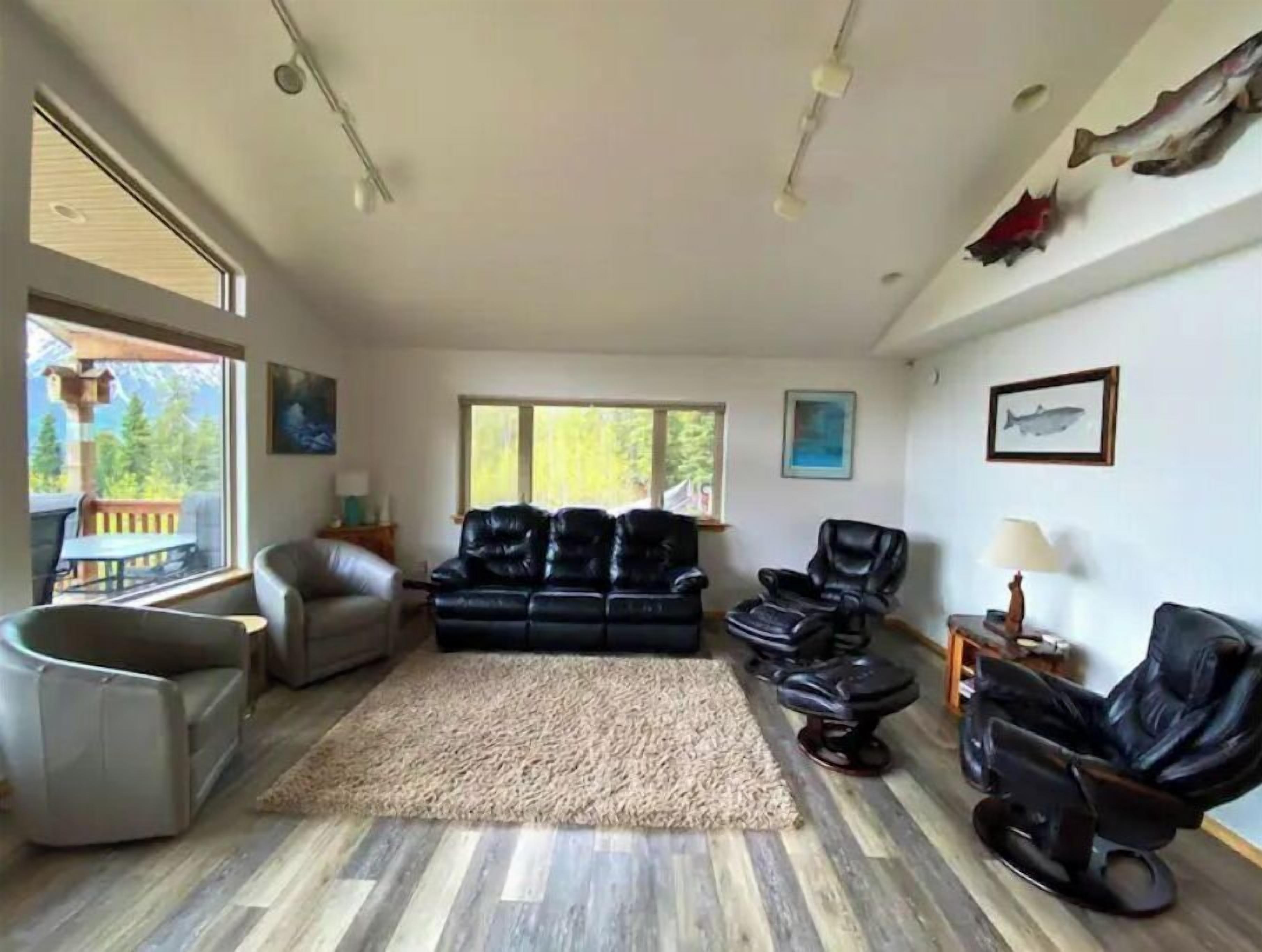 Un salón espacioso con un ventanal grande, un sofá de cuero negro, un sillón gris y suelo de madera.