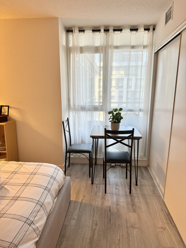 Une petite pièce comprenant un lit, une table avec deux chaises, une plante en pot et une fenêtre ornée de rideaux légers.