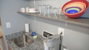 A kitchen with a granite countertop, stainless steel sink, and a coffee maker. There are shelves above holding glassware and plates. - Cute & Cozy North End Studio (Detroit)