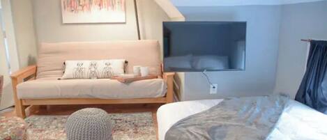 A bedroom with a sofa bed, a flat-screen TV mounted on the wall, a round ottoman, and a bed with a grey blanket.
