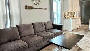 Un salón con sofá modular, una mesa de centro, suelo de madera, una zona de cocina con armarios blancos y una ventana con cortinas.