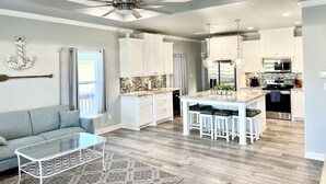 Una cocina moderna con armarios blancos, una isla central con taburetes de bar, un ventilador de techo y un comedor con mesa de vidrio.