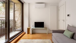 Un salón moderno con un televisor de pantalla plana sobre un mueble bajo, un sofá gris, suelo de madera y un amplio ventanal con vistas a paredes de piedra.
