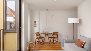 A modern living room with a grey sofa, a wooden dining table with two chairs, a white lamp, and a view of a red-roofed building outside. - JOIVY Stunning 1-bed flat with balcony (Porto)