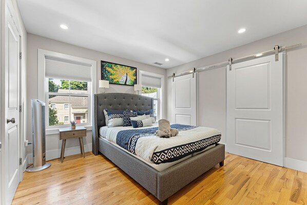 A bedroom with a large bed, a bedside table, a standing heater, and a window with blinds.