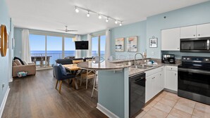 A modern kitchen with a central island, a dining area with a view, and a living room with a sofa and TV. - Beachfront Wrap Balcony & Views! Shores of Panama (Panama City Beach)