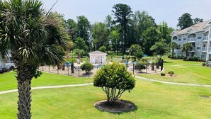 A well-maintained lawn with a small tree and a palm tree, surrounded by a fence and a building with multiple balconies. - Super Comfortable condo in center of Myrtle Beach (Myrtle Beach)