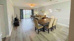 A spacious living room with a wooden dining table, beige chairs, a sofa, and a TV. - Super Comfortable condo in center of Myrtle Beach (Myrtle Beach)