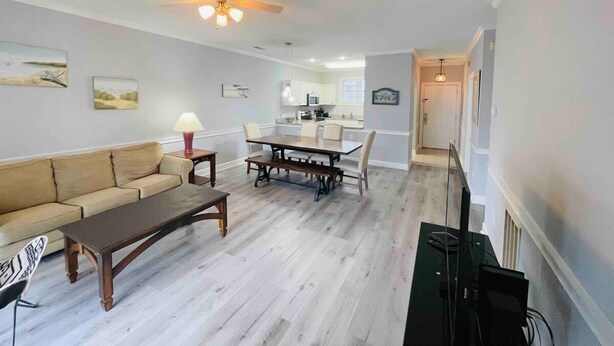 A living room with a sofa, coffee table, dining table, and chairs. There is a ceiling fan, a TV, and a kitchen in the background. - Super Comfortable condo in center of Myrtle Beach (Myrtle Beach)
