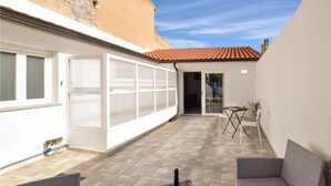 Un patio cubierto con suelo de baldosas, una puerta de cristal que da a un espacio interior, una mesa con sillas y una botella de champán sobre la mesa.
