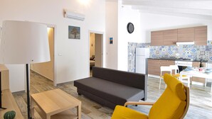Una sala de estar moderna con un sofá, un sillón amarillo, una mesa de centro de madera y una mesita con una lámpara. Hay una zona de cocina con baldosas estampadas y armarios de madera.