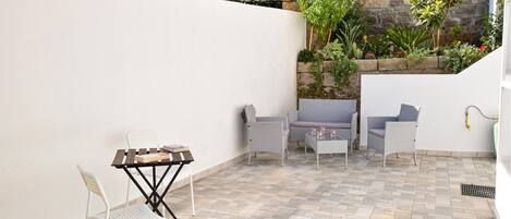 A patio with a white wall, a stone wall, and a tiled floor. There are two white chairs, a small table, and a white folding chair.