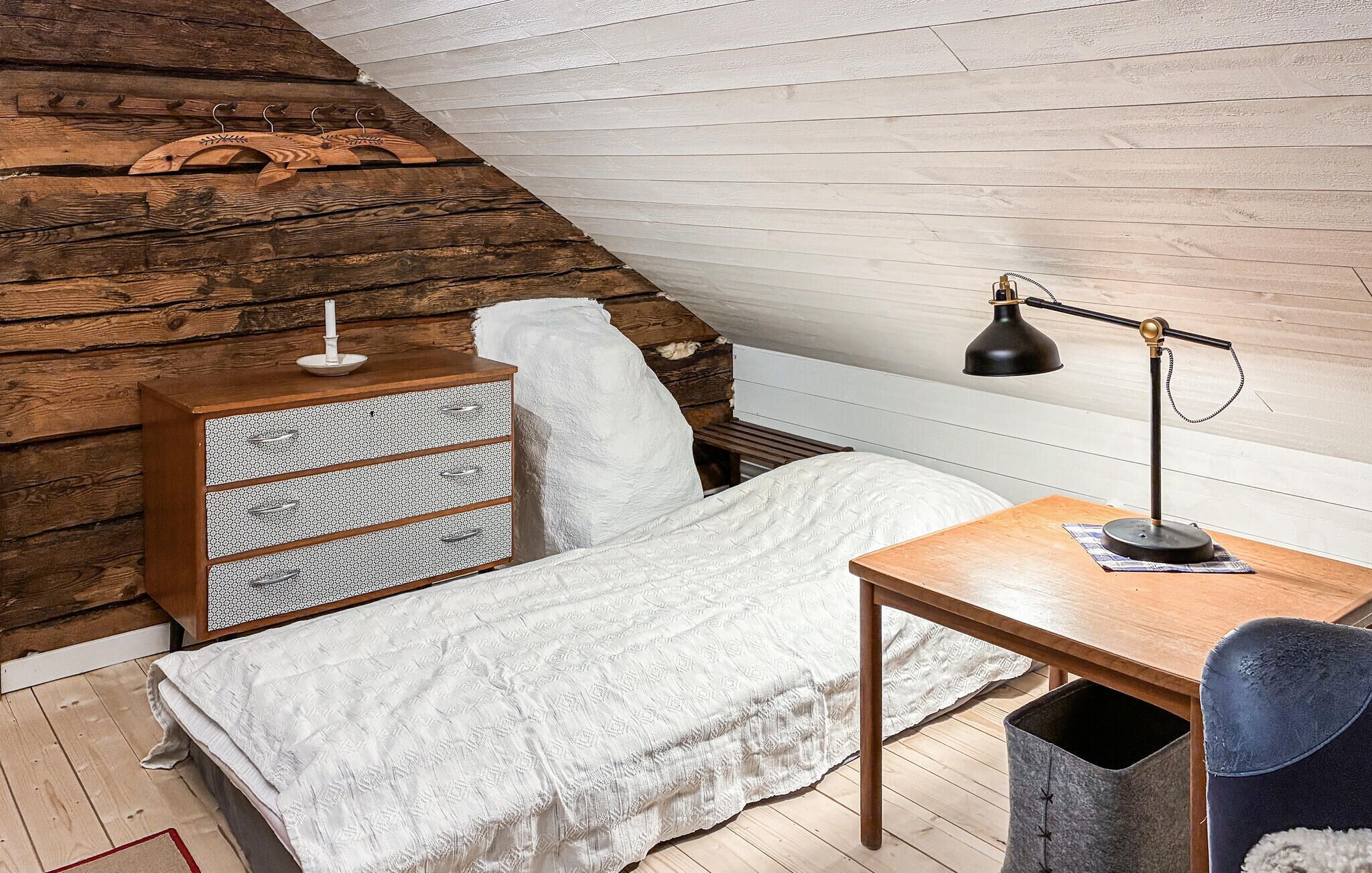A loft bedroom with a sloped ceiling, a wooden dresser, a bed with a white blanket, a desk with a lamp, and a chair.