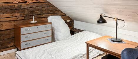 A loft bedroom with a sloped ceiling, a wooden dresser, a bed with a white blanket, a desk with a lamp, and a chair.