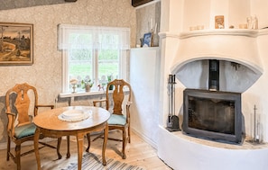 A cozy room with a fireplace, a dining table with chairs, a window with a view of greenery, and a framed picture on the wall.