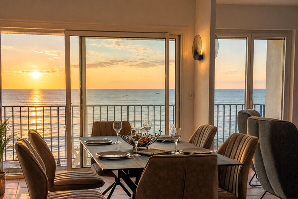 A dining area with a table set for two, overlooking the sea, with a balcony and a view of the sunset.