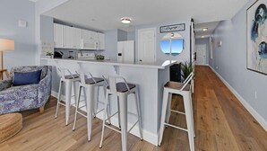 A modern kitchen with white cabinets, a bar area with stools, and a view of the ocean through a window. - BeachFront-Ocean View at Majestic Beach Resort #906 (Panama City)