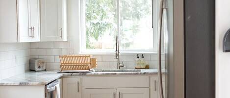 A kitchen with white cabinets, a stainless steel refrigerator, a window with blinds, and a wooden floor.