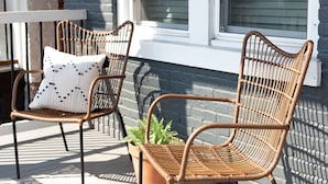 Zwei Korbstühle mit Kissen, auf einer Holzterrasse platziert, mit einem Blumentopf dazwischen.