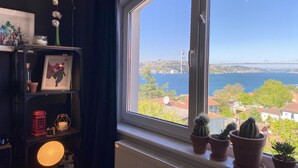 Een kamer met een groot raam dat uitkijkt op een wateroppervlak en een brug, een radiator, een plank met diverse spullen en een brandende lamp.